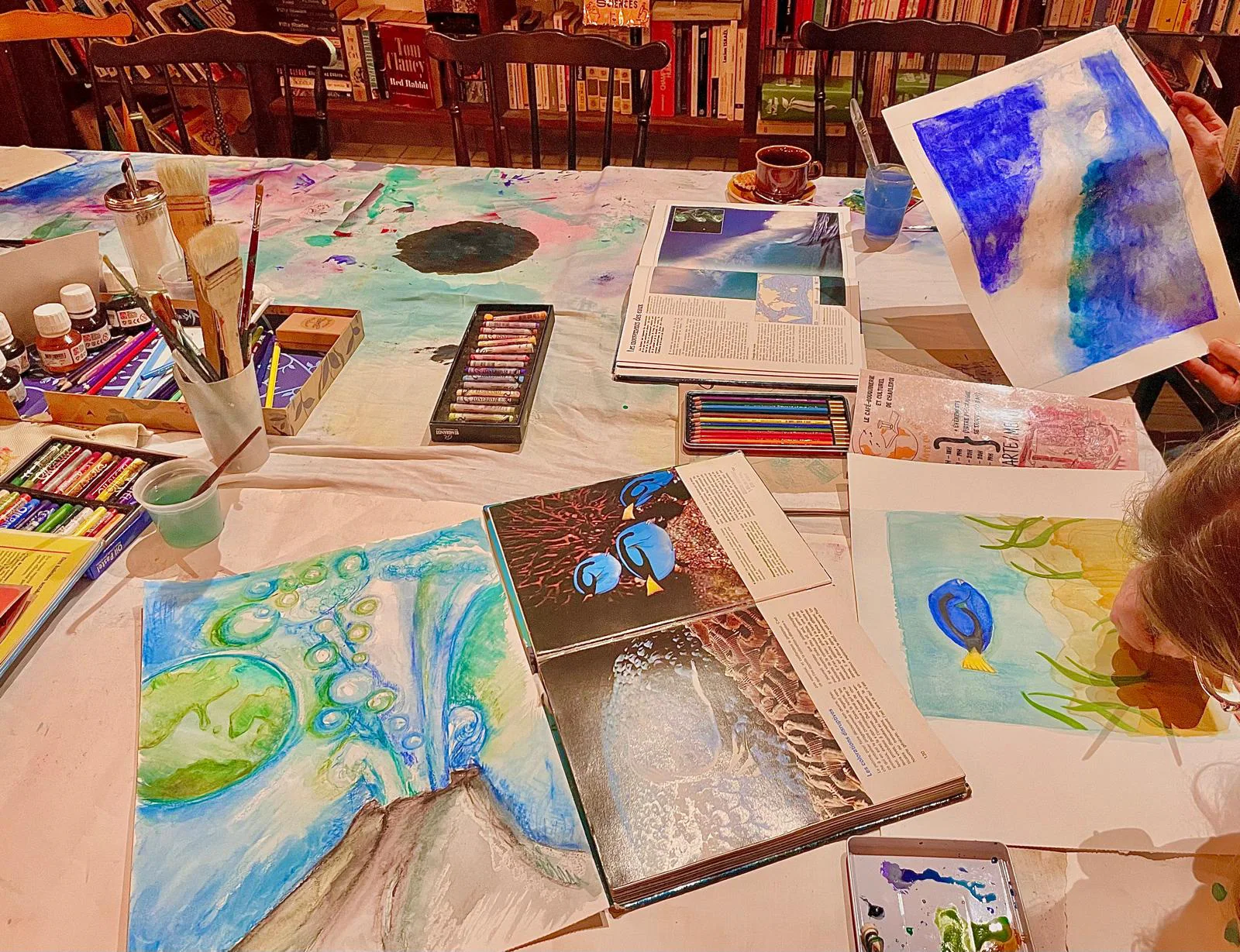 Table d'atelier couverte d'aquarelles marines, de pastels et de pinceaux.