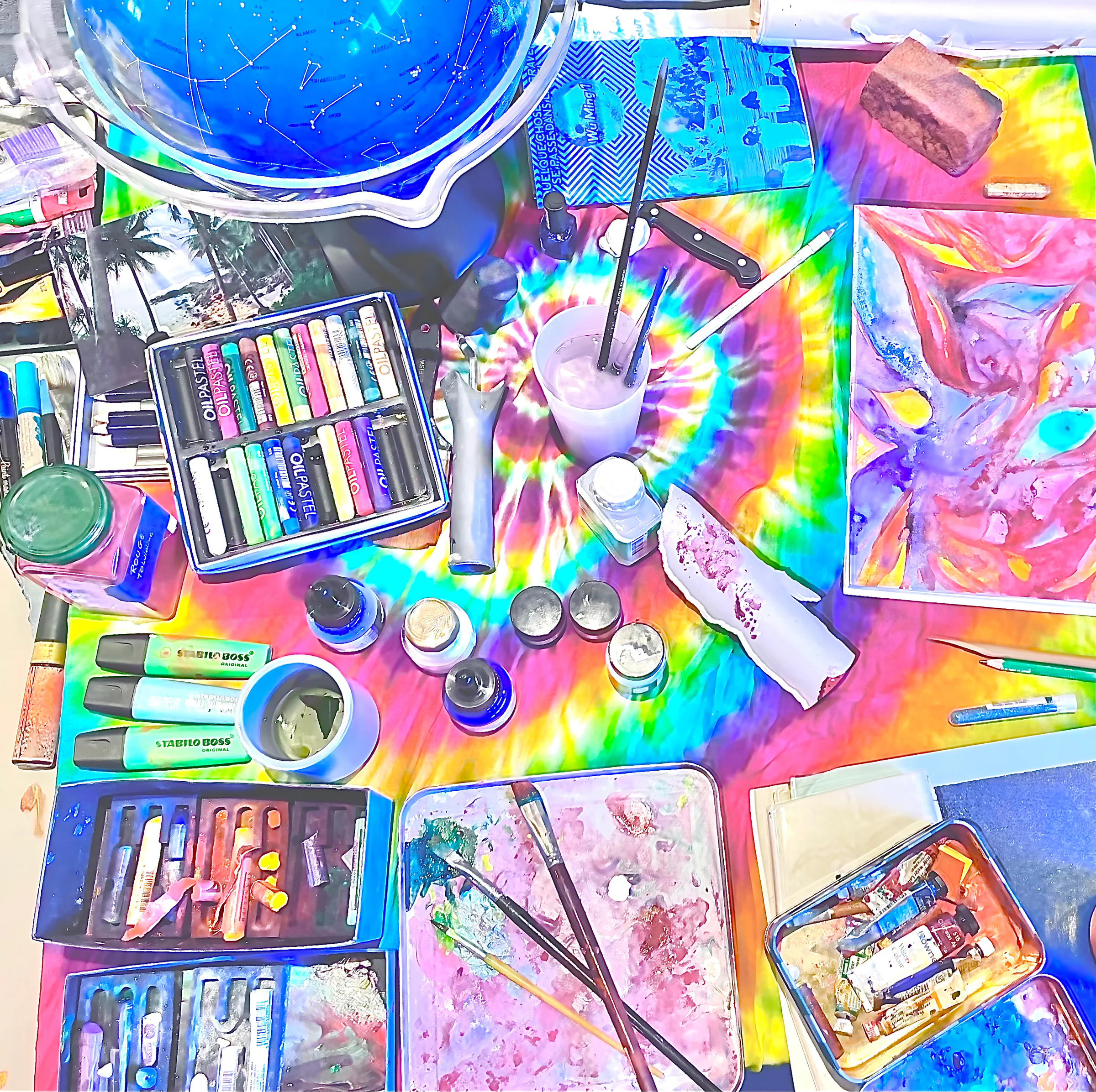 Vue du dessus d'une table d'atelier, pastels, godets et papiers aux couleurs vives.